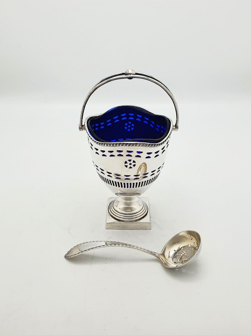 A silver-plated pierced pedestal sugar basket with a swing handle, cobalt blue glass liner, and accompanying sifter spoon.