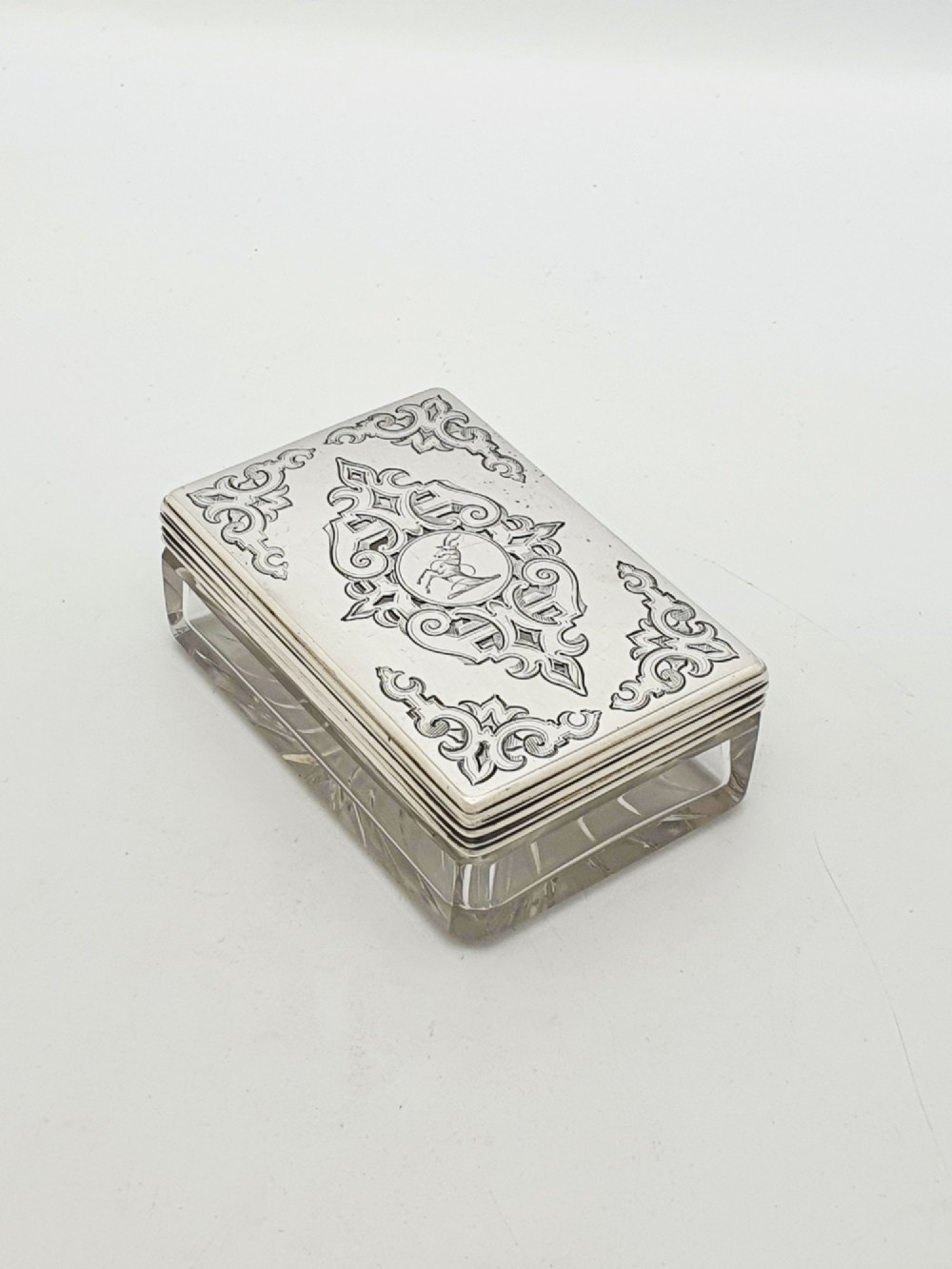 A rectangular cut glass trinket box featuring a sterling silver lid engraved with elaborate scrollwork and a central stag crest.