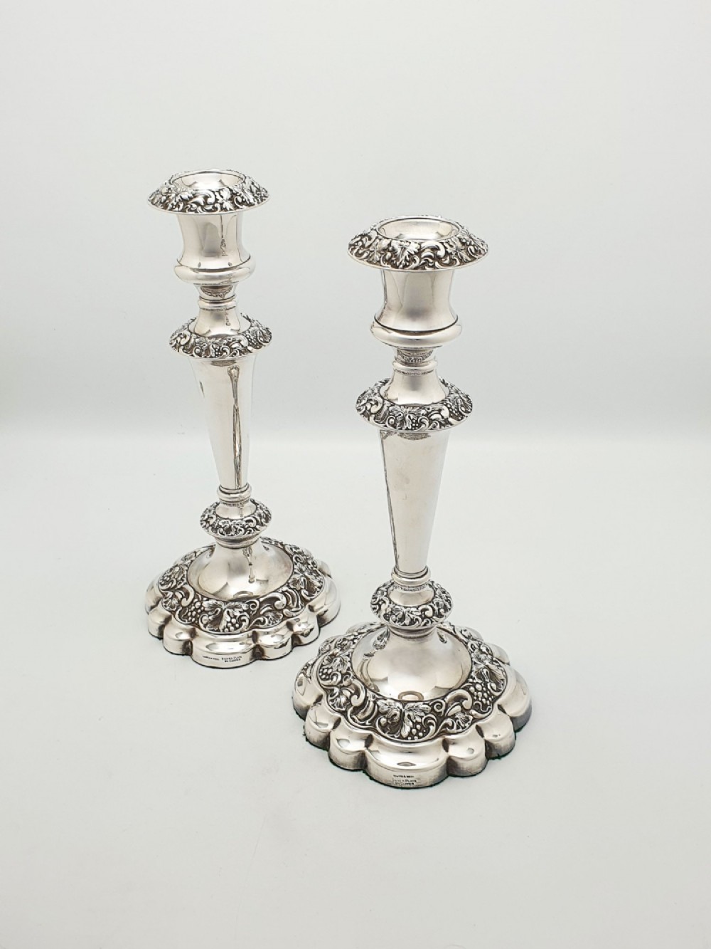 A pair of polished silver-plated candlesticks featuring ornate foliate repousse borders on scalloped bases.