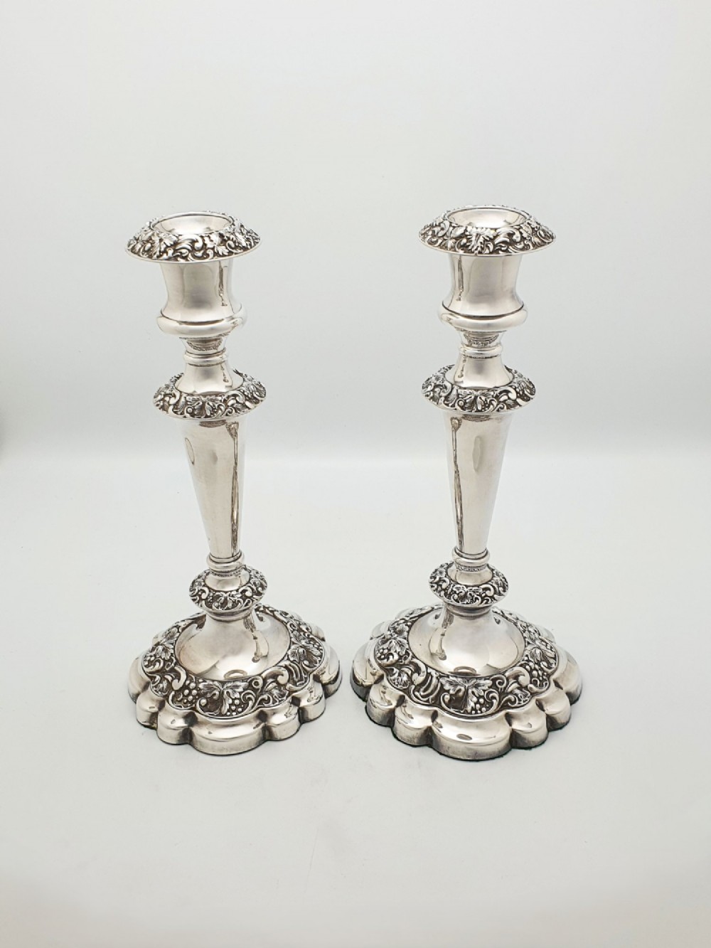 A pair of polished silver-plated candlesticks featuring ornate foliate repousse borders on scalloped bases. View V