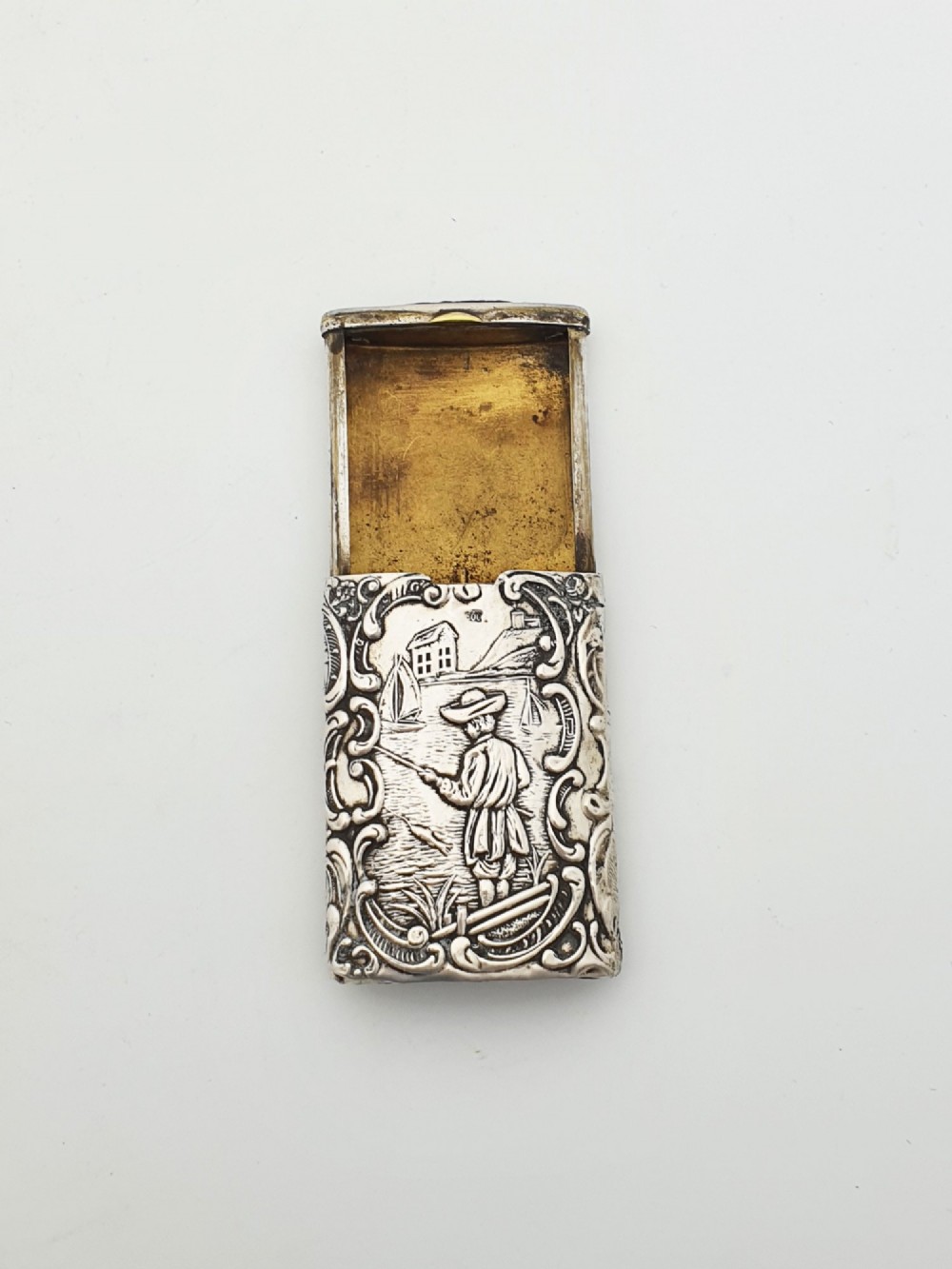 A rectangular silver vesta case with detailed embossed scrollwork around a scenic relief of a figure by water, featuring a sliding gilt interior compartment.