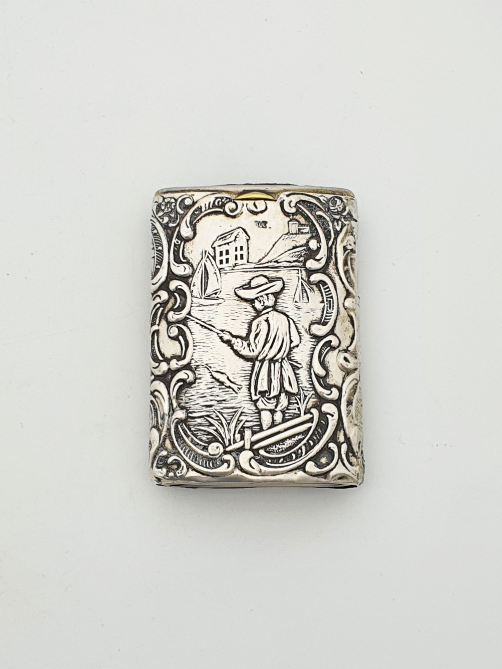 A rectangular silver vesta case with detailed embossed scrollwork around a scenic relief of a figure by water, featuring a sliding gilt interior compartment. View II