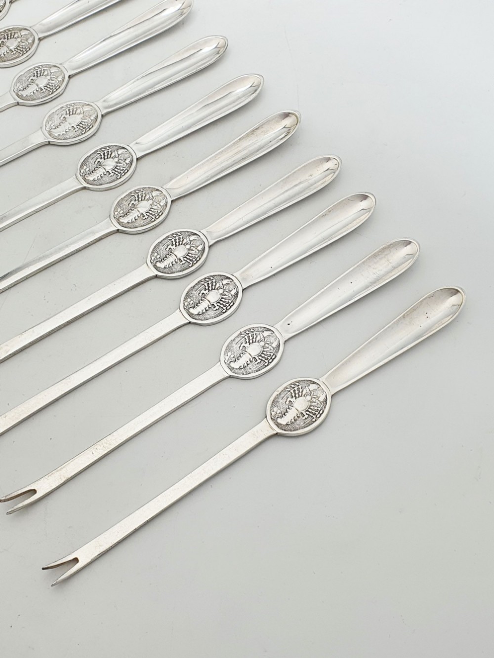 A set of twelve silver-plated lobster picks featuring embossed lobster medallions on the handles and narrow two-pronged tips, arranged neatly in a diagonal row. View II