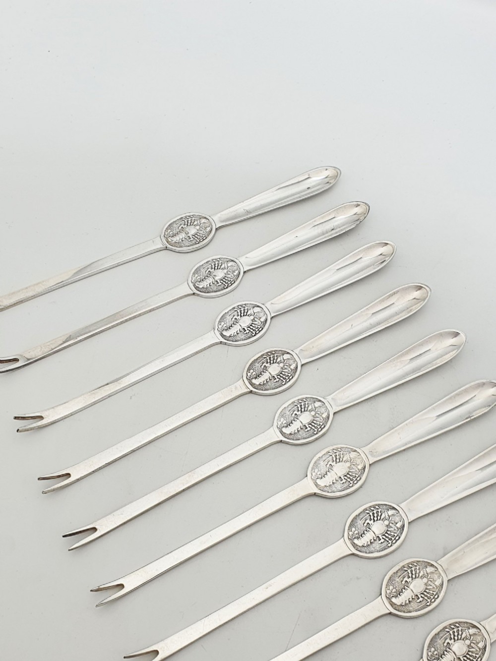 A set of twelve silver-plated lobster picks featuring embossed lobster medallions on the handles and narrow two-pronged tips, arranged neatly in a diagonal row. View III