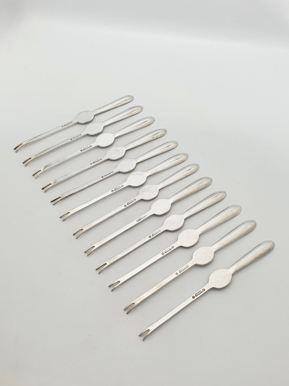 A set of twelve silver-plated lobster picks featuring embossed lobster medallions on the handles and narrow two-pronged tips, arranged neatly in a diagonal row. View IV
