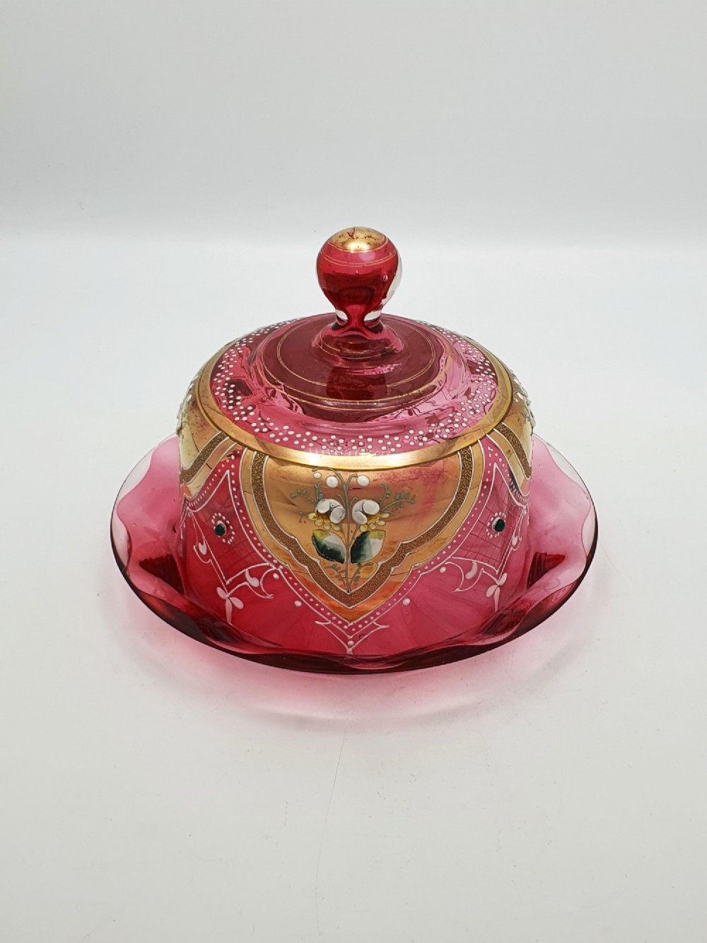 An antique cranberry red glass covered cheese dish featuring gold filigree detailing, floral enamel panels, and a rounded glass knob finial.
