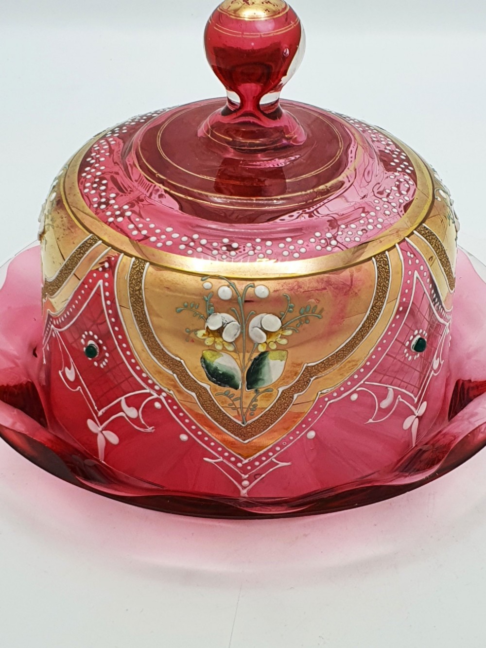 An antique cranberry red glass covered cheese dish featuring gold filigree detailing, floral enamel panels, and a rounded glass knob finial. View II