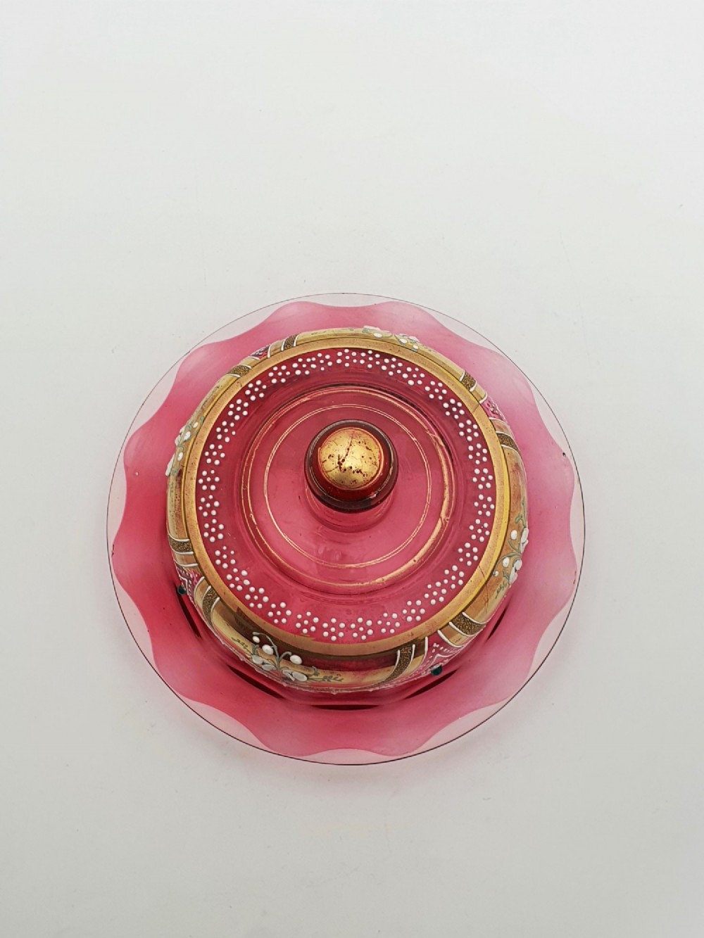 An antique cranberry red glass covered cheese dish featuring gold filigree detailing, floral enamel panels, and a rounded glass knob finial. View III