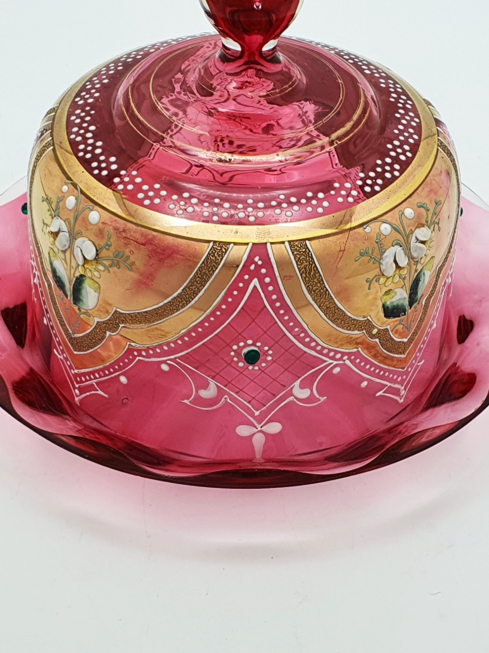 An antique cranberry red glass covered cheese dish featuring gold filigree detailing, floral enamel panels, and a rounded glass knob finial. View IV