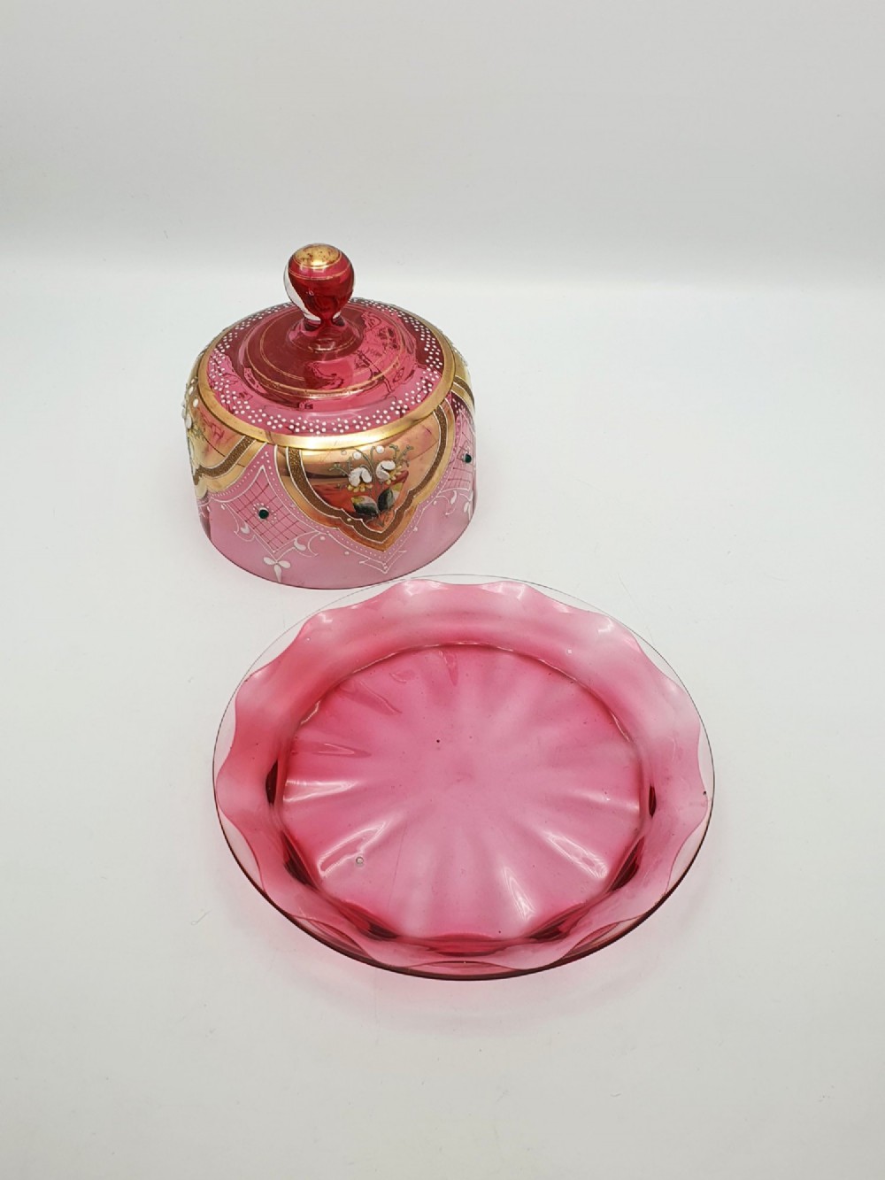 An antique cranberry red glass covered cheese dish featuring gold filigree detailing, floral enamel panels, and a rounded glass knob finial. View V
