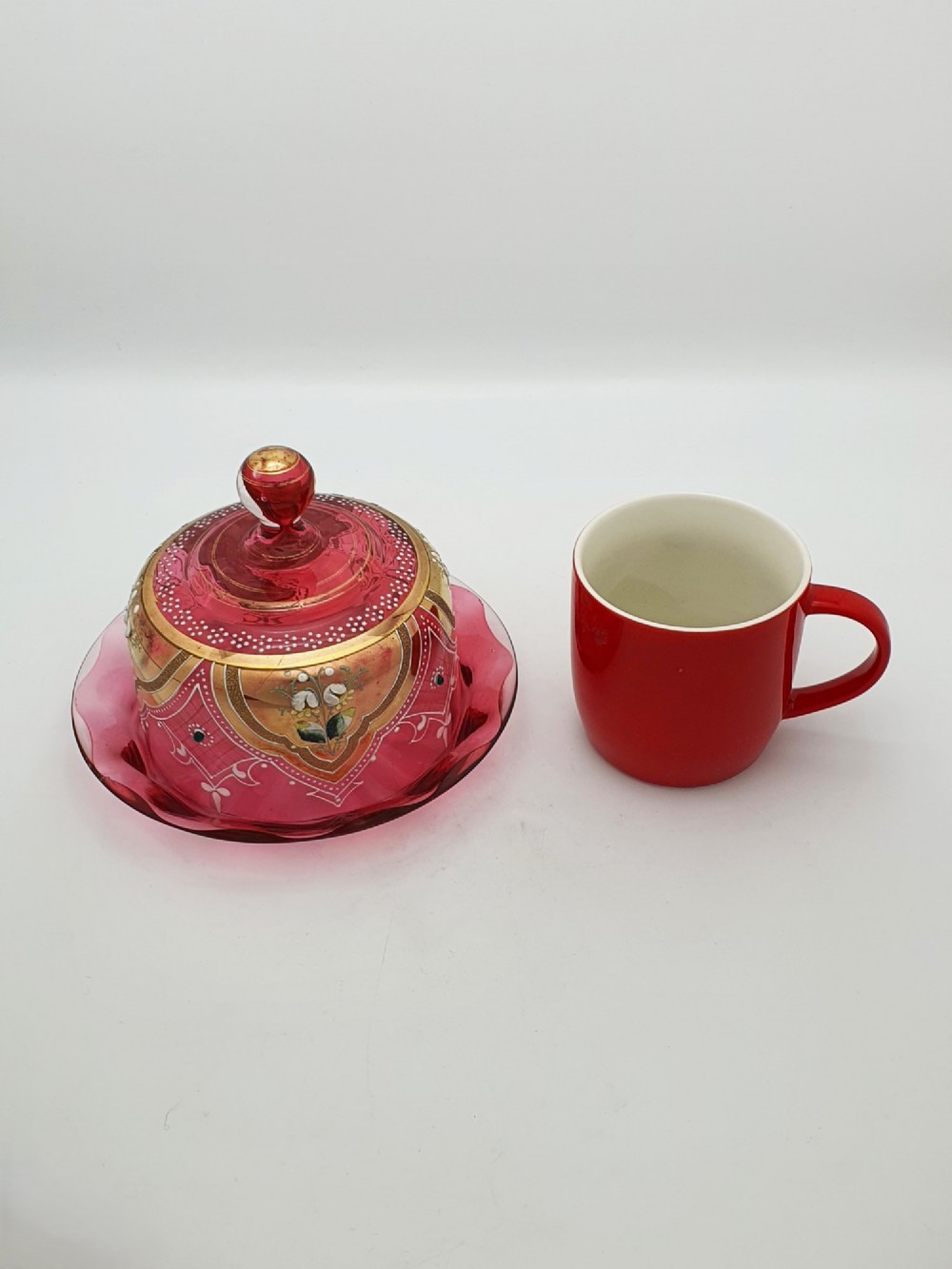 An antique cranberry red glass covered cheese dish featuring gold filigree detailing, floral enamel panels, and a rounded glass knob finial. View VII