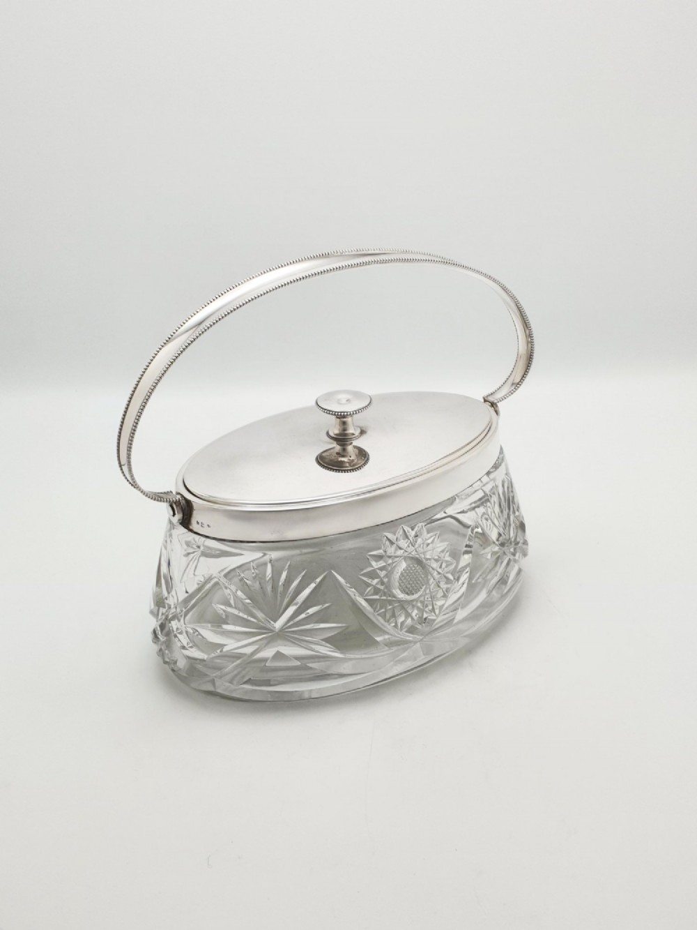 An oval cut glass biscuit box featuring a starburst motif, fitted with a silver-plated lid, central finial, and a beaded swing handle.