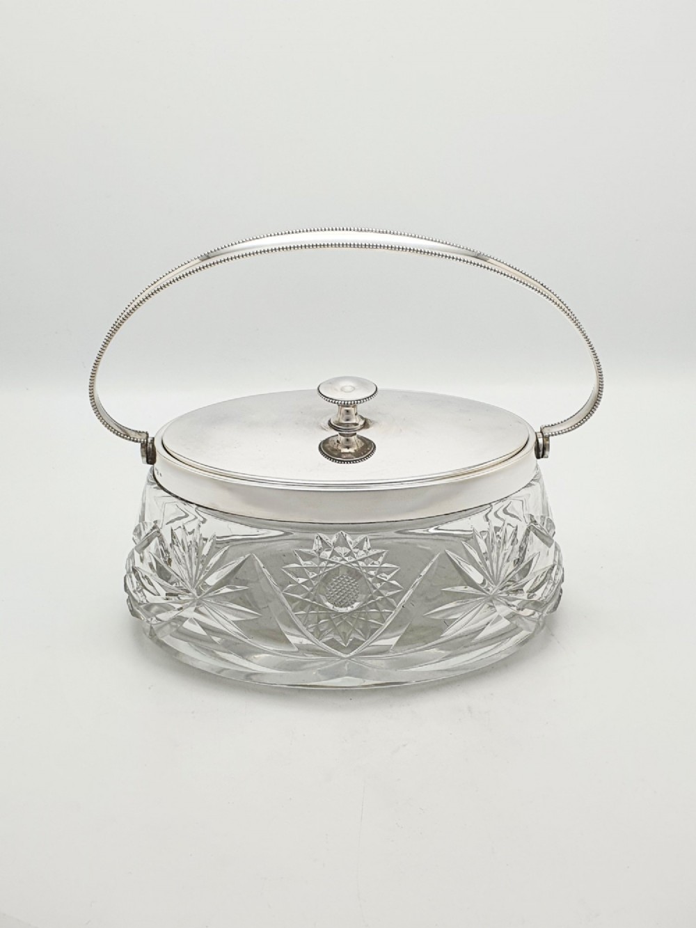 An oval cut glass biscuit box featuring a starburst motif, fitted with a silver-plated lid, central finial, and a beaded swing handle. View II