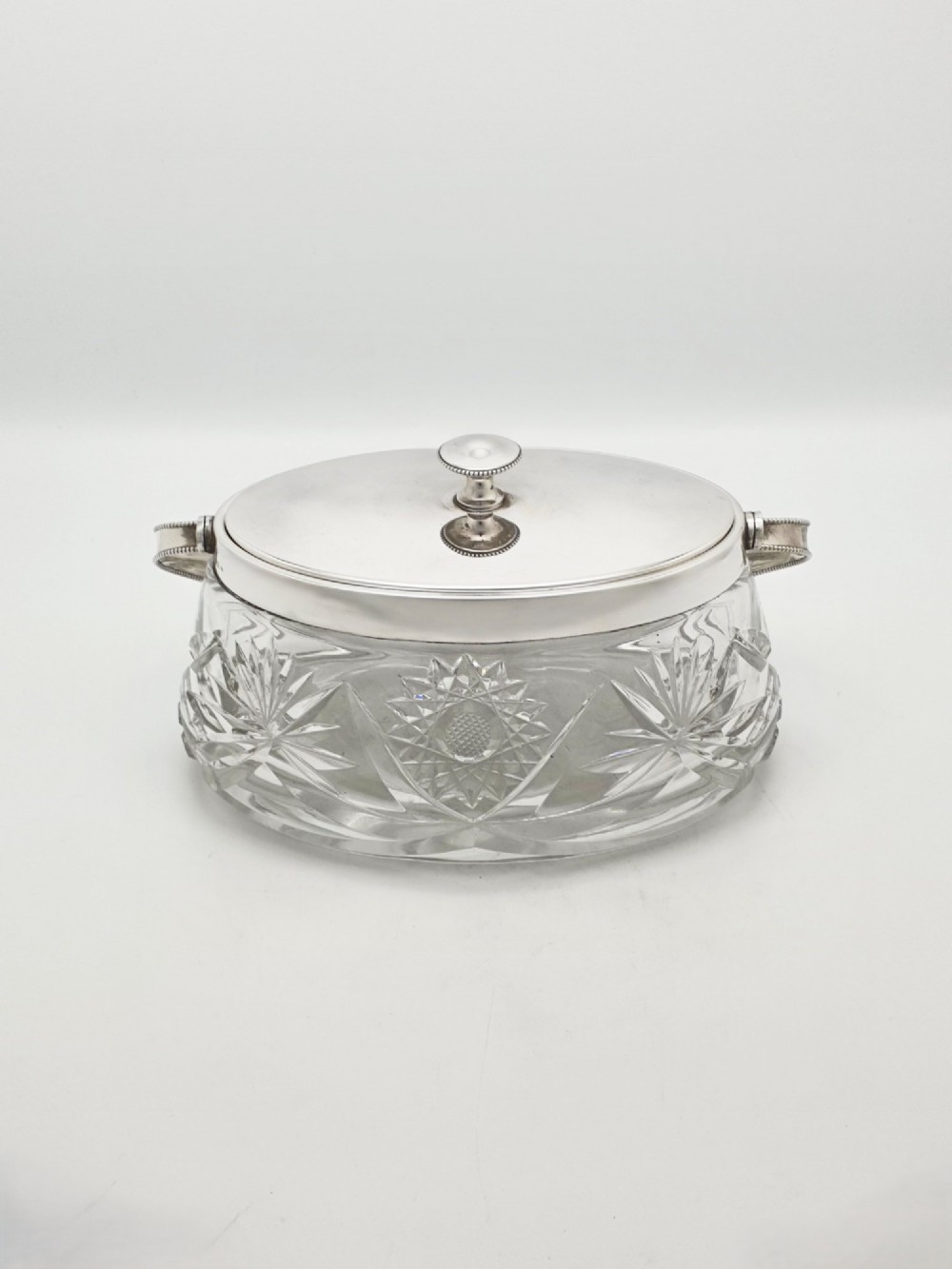 An oval cut glass biscuit box featuring a starburst motif, fitted with a silver-plated lid, central finial, and a beaded swing handle. View III