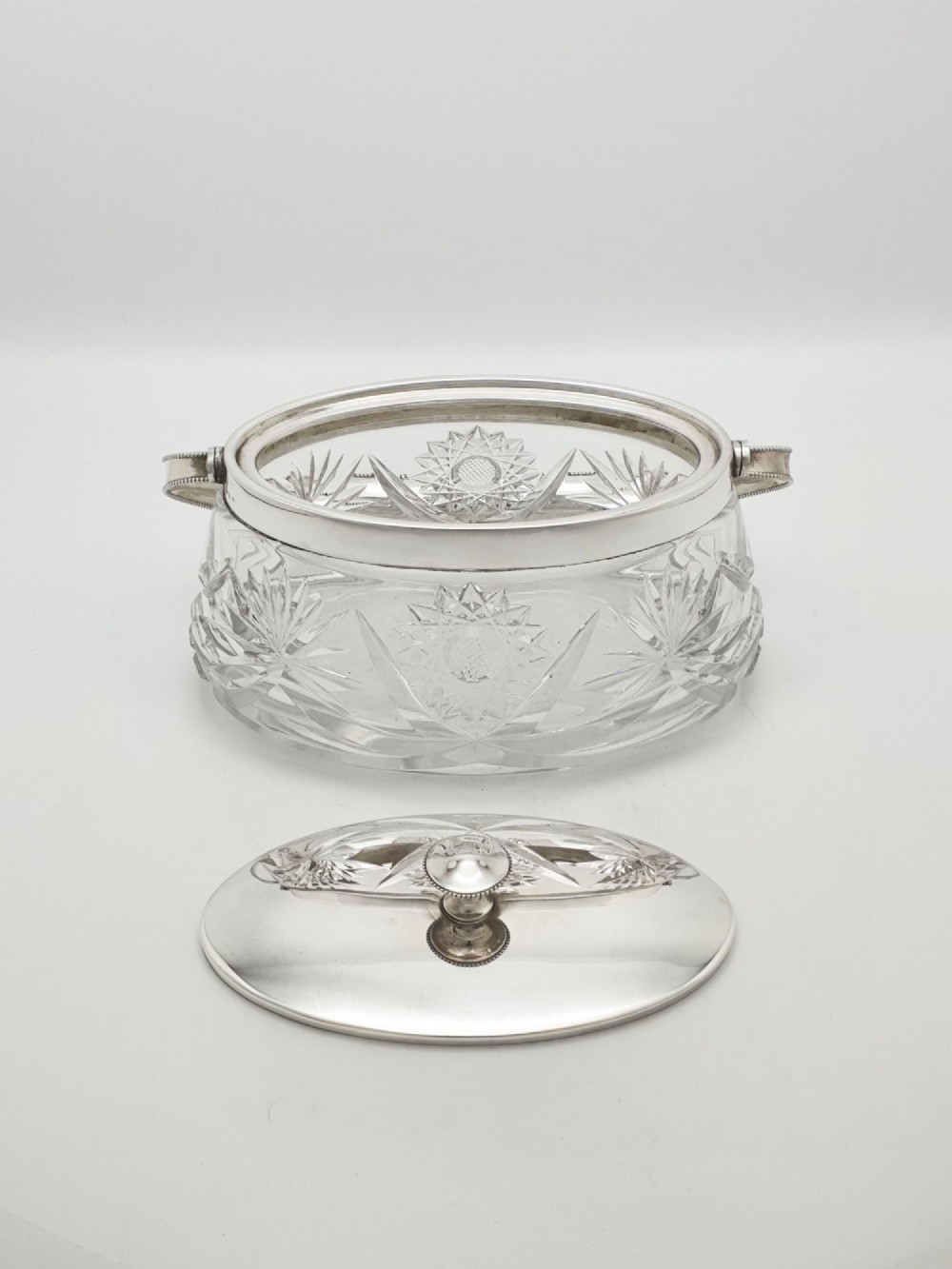 An oval cut glass biscuit box featuring a starburst motif, fitted with a silver-plated lid, central finial, and a beaded swing handle. View IV
