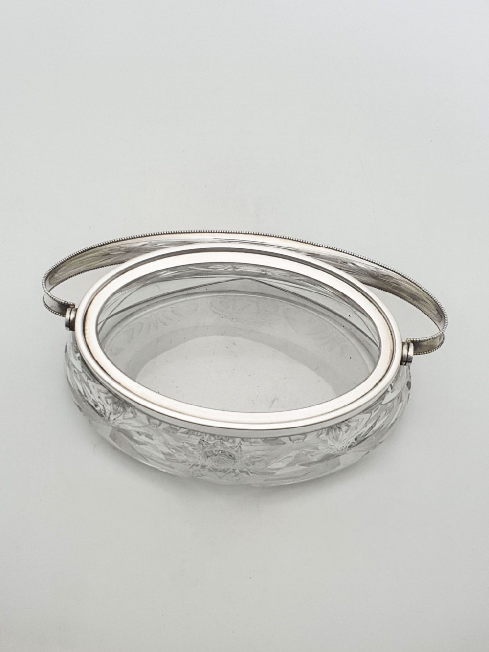 An oval cut glass biscuit box featuring a starburst motif, fitted with a silver-plated lid, central finial, and a beaded swing handle. View V