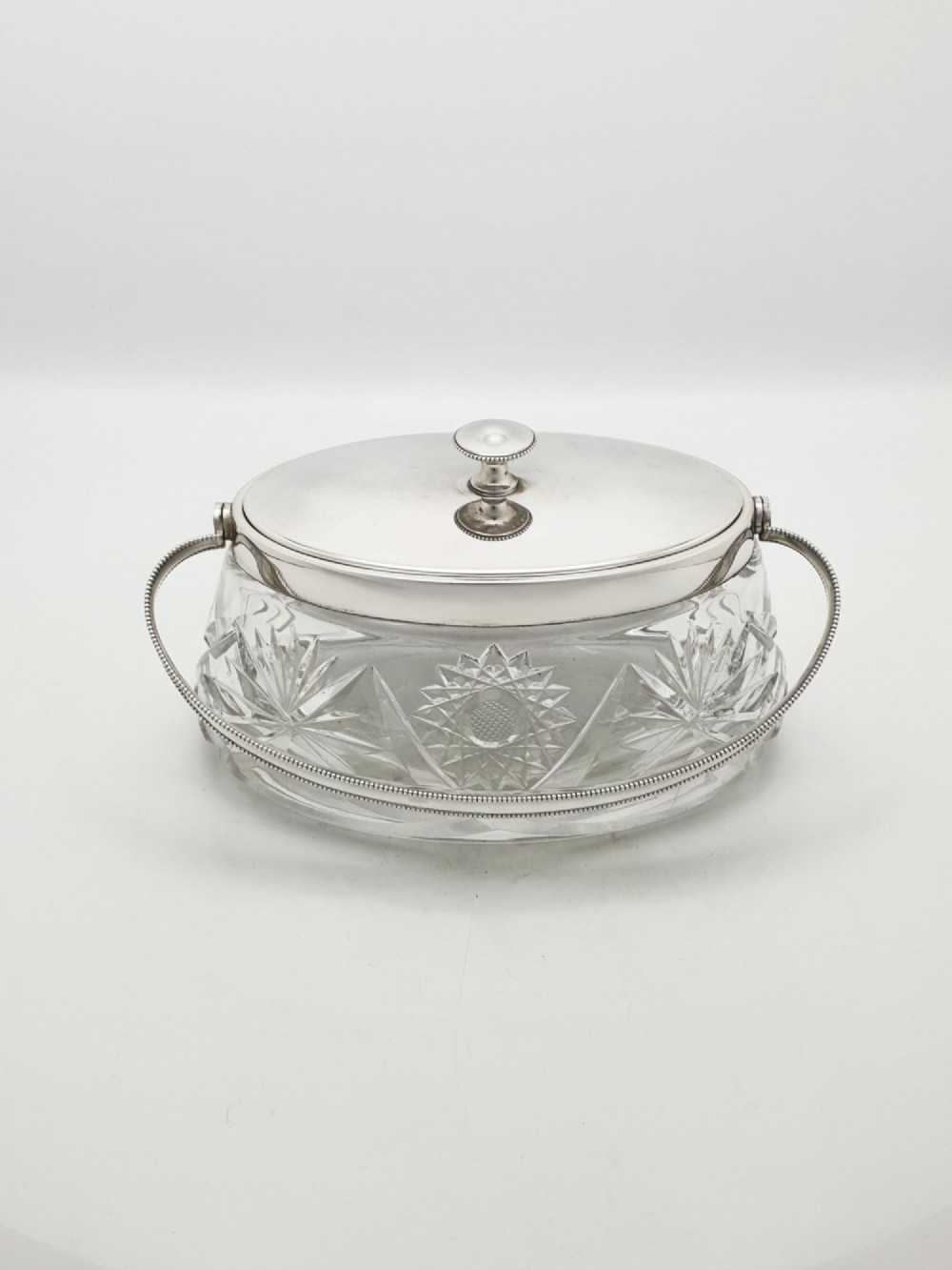 An oval cut glass biscuit box featuring a starburst motif, fitted with a silver-plated lid, central finial, and a beaded swing handle. View VI