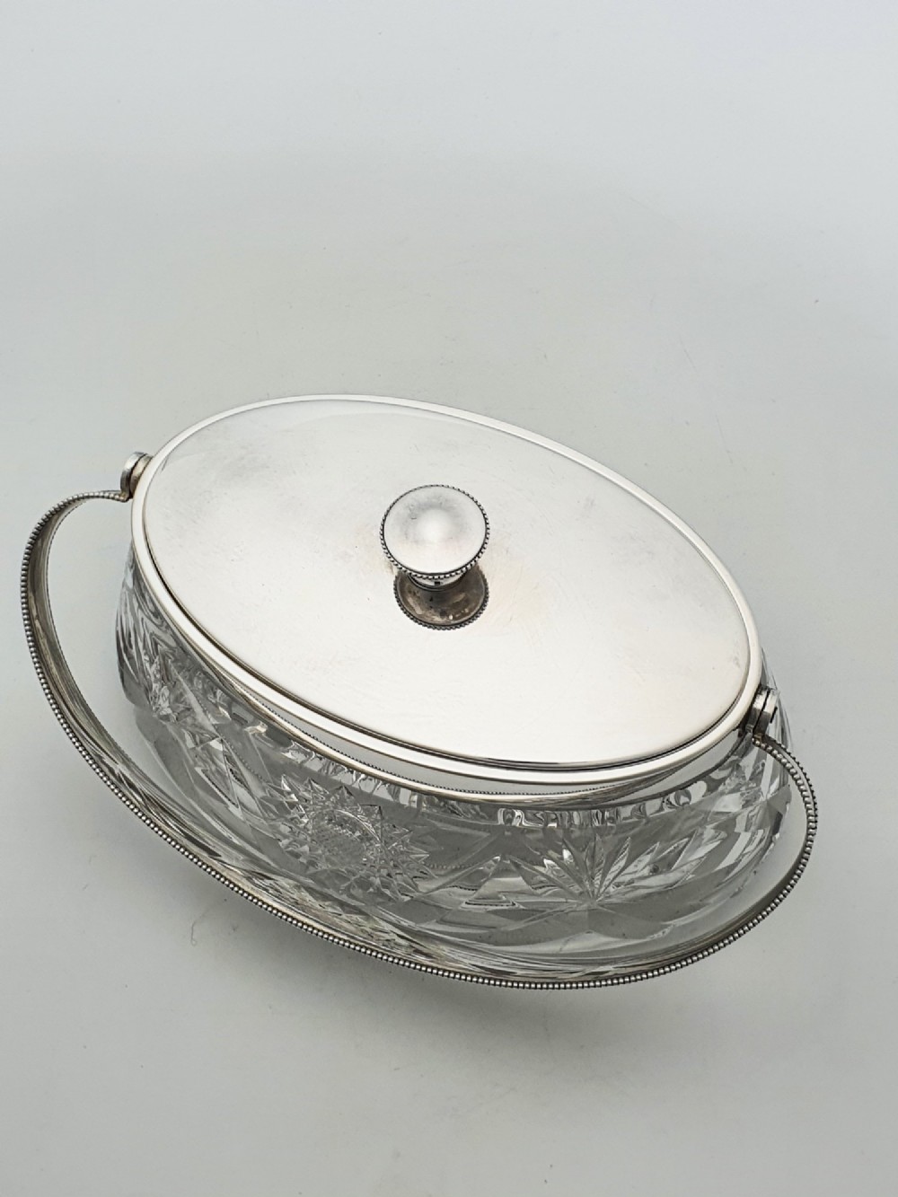 An oval cut glass biscuit box featuring a starburst motif, fitted with a silver-plated lid, central finial, and a beaded swing handle. View VII