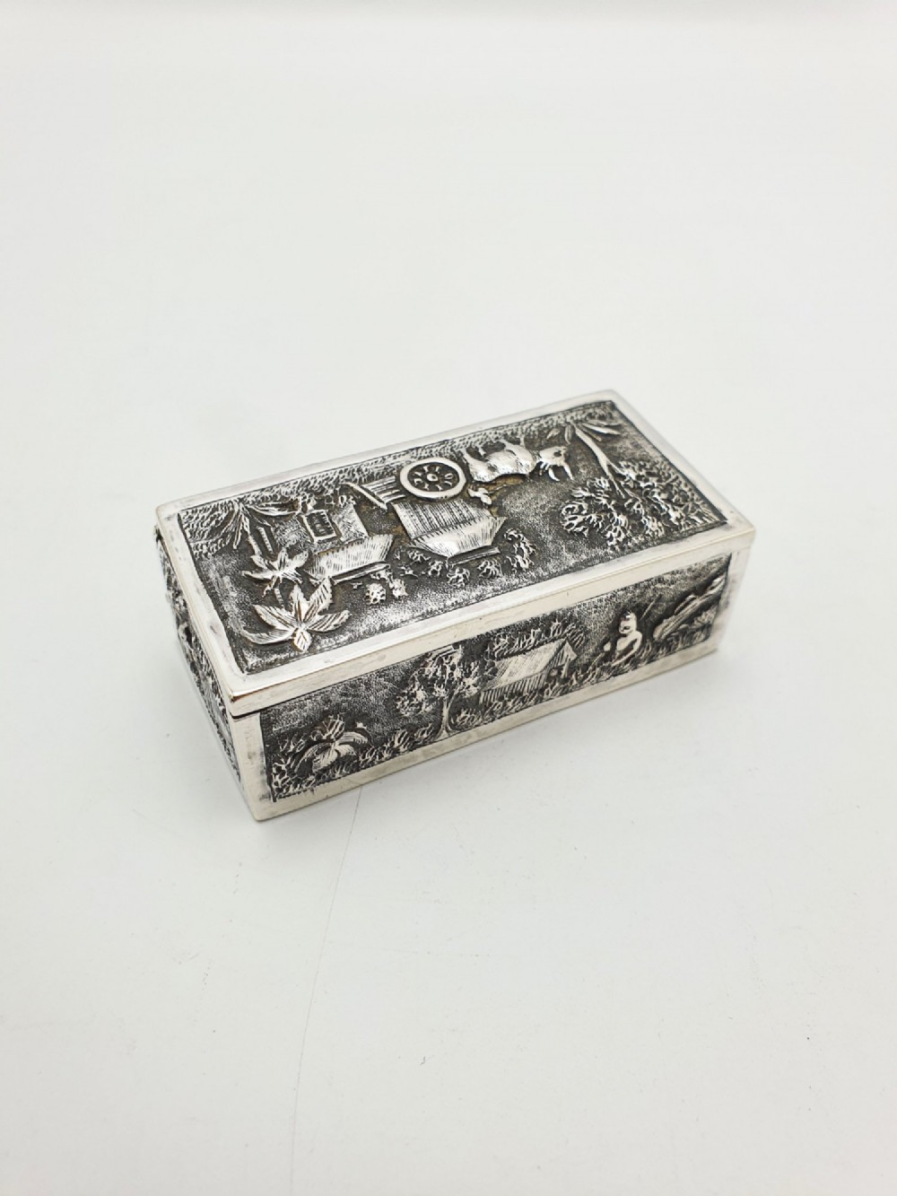 A rectangular antique silver trinket box featuring detailed repousse relief depicting a village scene with huts and palm trees.