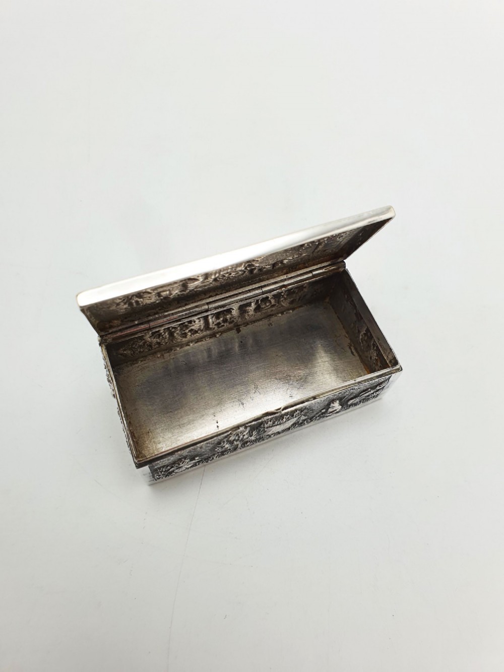 A rectangular antique silver trinket box featuring detailed repousse relief depicting a village scene with huts and palm trees. View III
