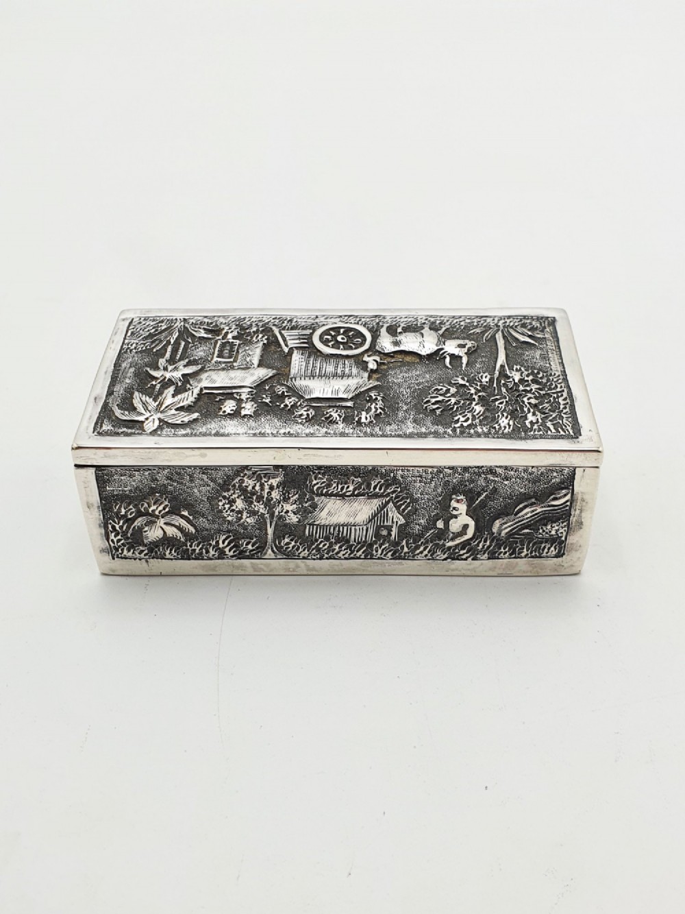 A rectangular antique silver trinket box featuring detailed repousse relief depicting a village scene with huts and palm trees. View IV