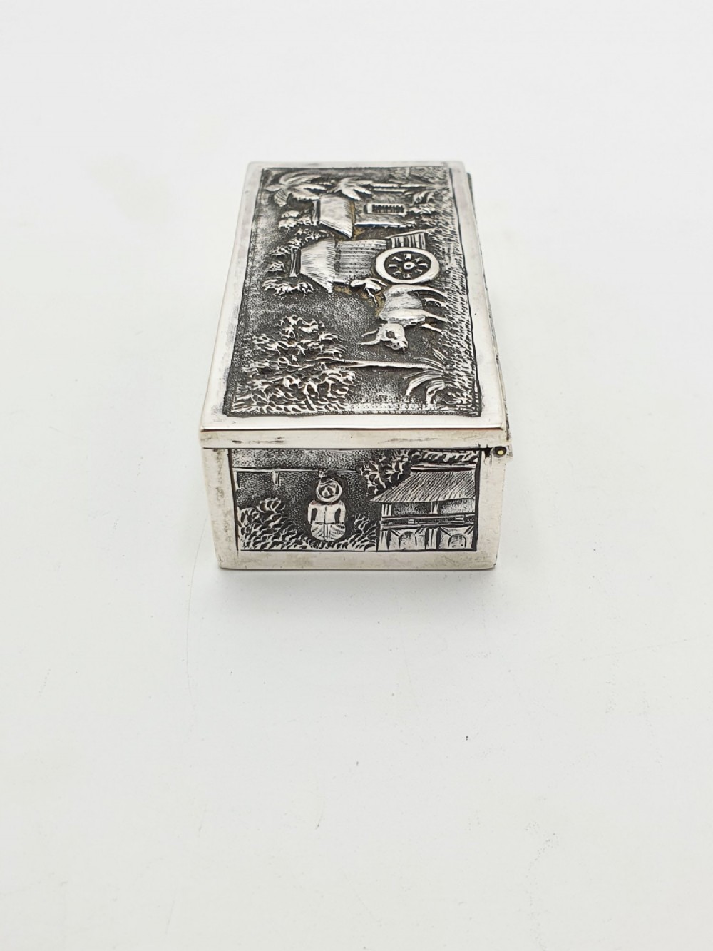 A rectangular antique silver trinket box featuring detailed repousse relief depicting a village scene with huts and palm trees. View V