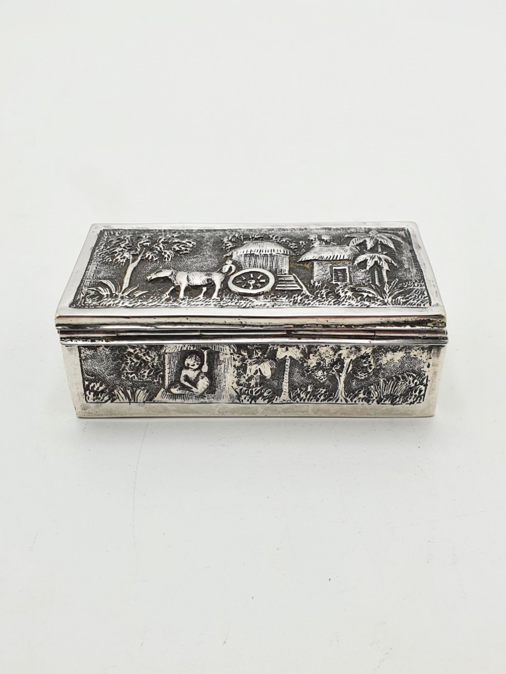 A rectangular antique silver trinket box featuring detailed repousse relief depicting a village scene with huts and palm trees. View VI
