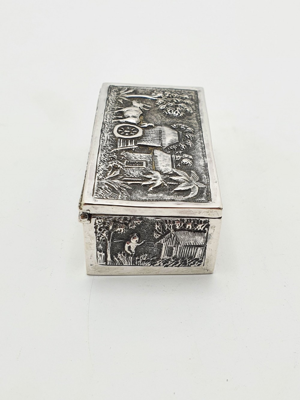 A rectangular antique silver trinket box featuring detailed repousse relief depicting a village scene with huts and palm trees. View VII