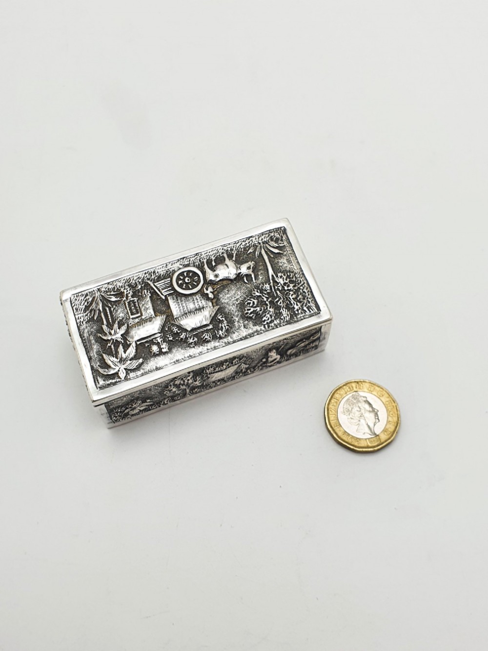 A rectangular antique silver trinket box featuring detailed repousse relief depicting a village scene with huts and palm trees. View IX
