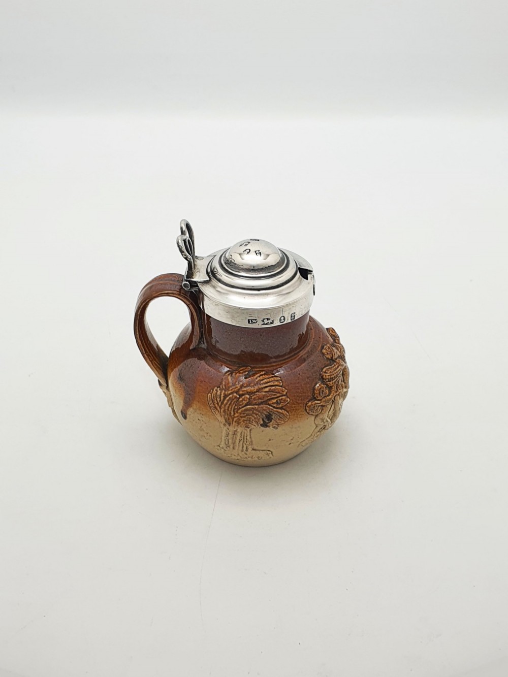 A small salt-glazed brown and beige stoneware jug featuring relief motifs and a hallmarked sterling silver hinged lid and collar.