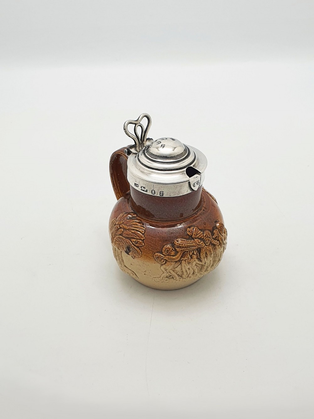 A small salt-glazed brown and beige stoneware jug featuring relief motifs and a hallmarked sterling silver hinged lid and collar. View II