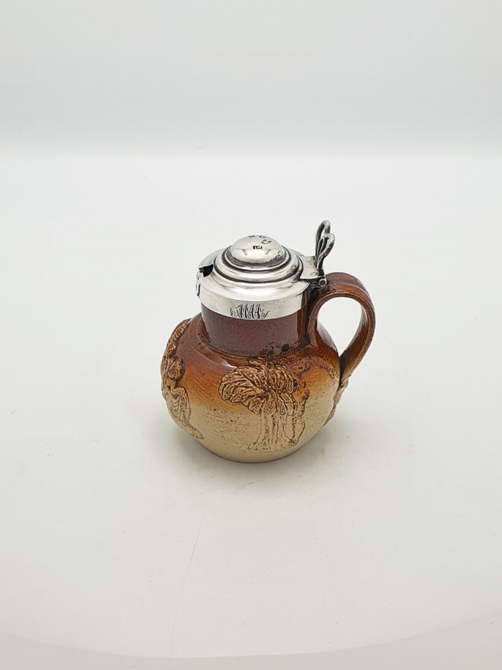 A small salt-glazed brown and beige stoneware jug featuring relief motifs and a hallmarked sterling silver hinged lid and collar. View V
