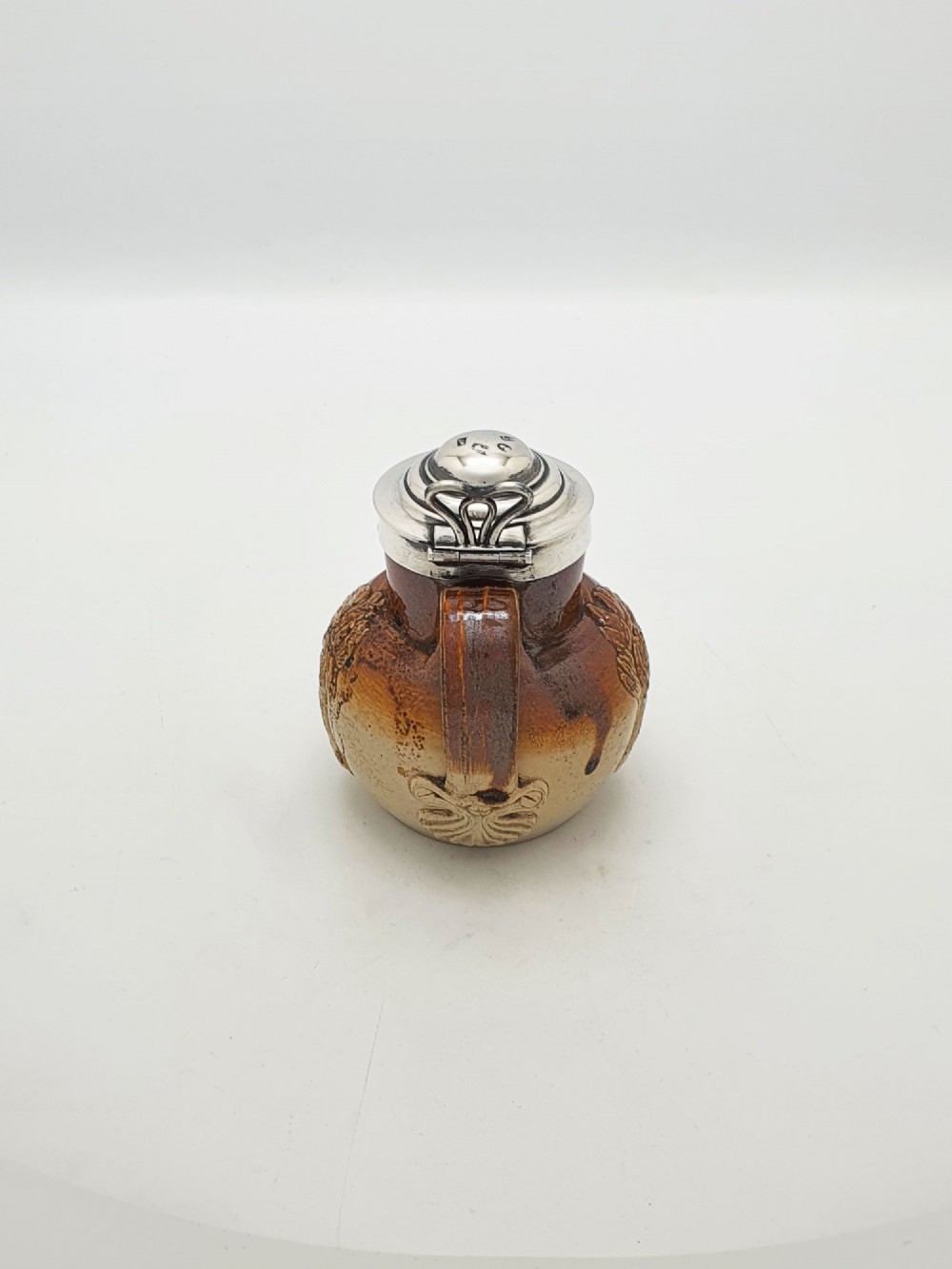 A small salt-glazed brown and beige stoneware jug featuring relief motifs and a hallmarked sterling silver hinged lid and collar. View VI