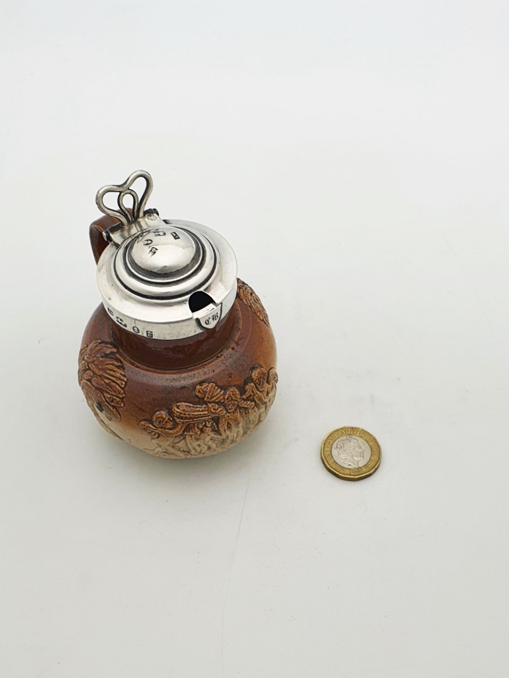 A small salt-glazed brown and beige stoneware jug featuring relief motifs and a hallmarked sterling silver hinged lid and collar. View X