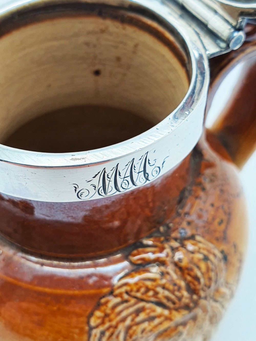 A small salt-glazed brown and beige stoneware jug featuring relief motifs and a hallmarked sterling silver hinged lid and collar. View XII