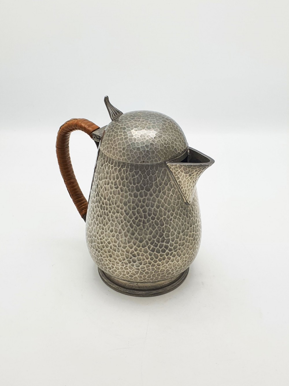 A planished pewter coffee pot featuring a domed lid, triangular spout, and a brown cane-wrapped handle.