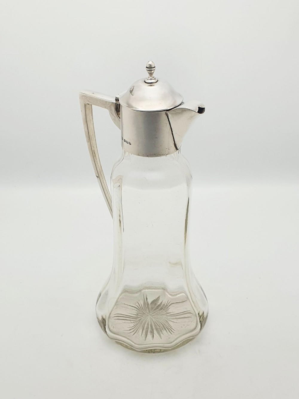 An Edwardian clear glass claret jug featuring a star-cut base, fluted body, angular silver handle, and sterling silver mounted spout with a domed lid.