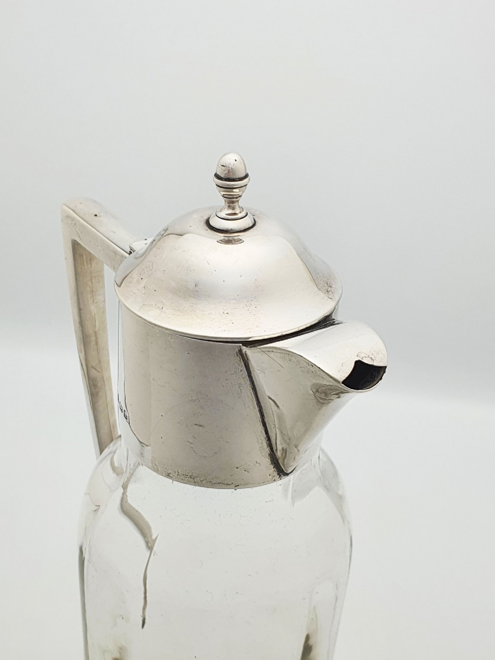 An Edwardian clear glass claret jug featuring a star-cut base, fluted body, angular silver handle, and sterling silver mounted spout with a domed lid. View IX