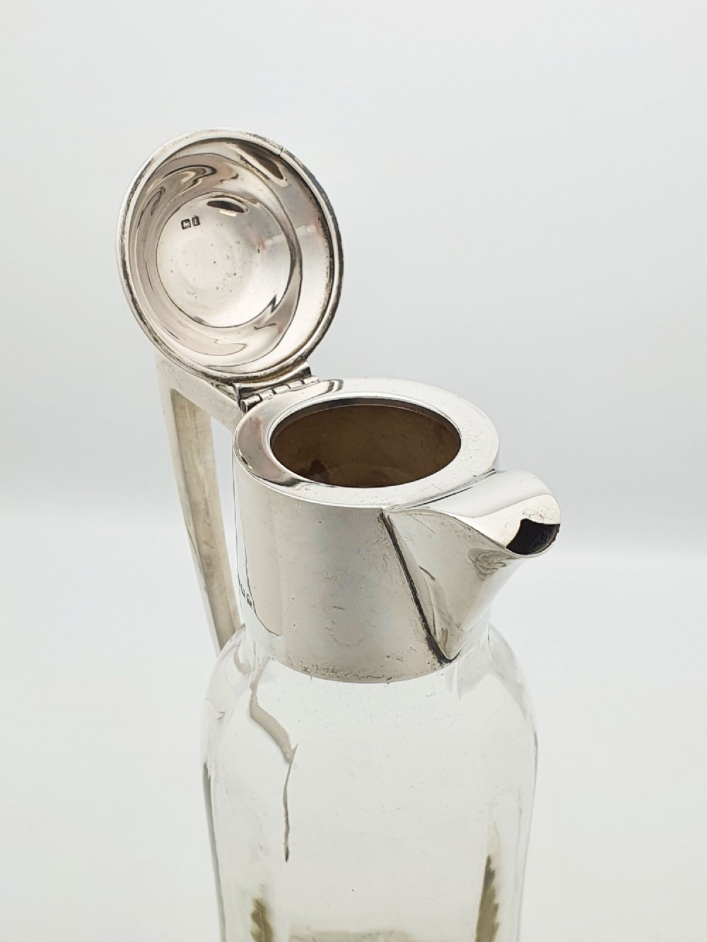 An Edwardian clear glass claret jug featuring a star-cut base, fluted body, angular silver handle, and sterling silver mounted spout with a domed lid. View X