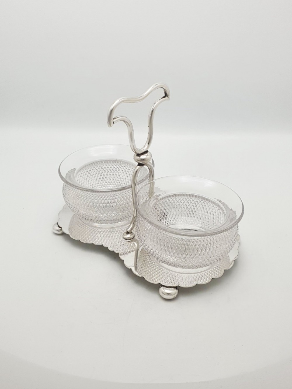 A silver-plated condiment stand featuring a central looped handle, ball feet, and two clear diamond-patterned glass bowls.