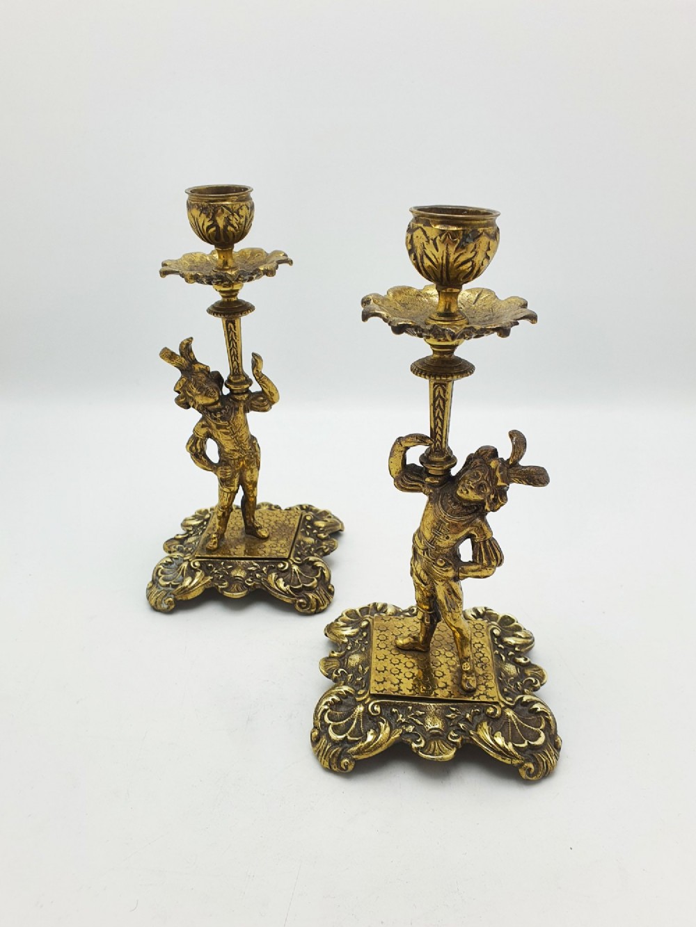 A pair of Victorian gilt brass candlesticks featuring sculpted figural stems raised on square ornamented bases with acanthus detail.