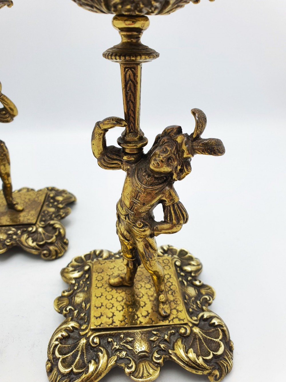 A pair of Victorian gilt brass candlesticks featuring sculpted figural stems raised on square ornamented bases with acanthus detail. View II