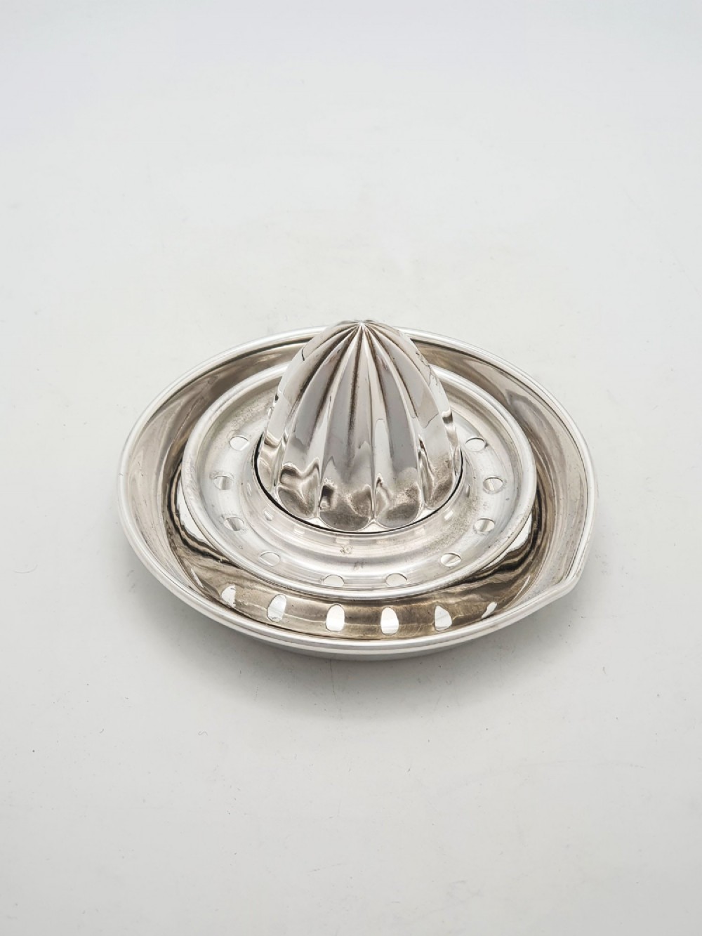 A silver-plated manual citrus squeezer featuring a central fluted reamer cone, a circular perforated strainer ring, and a shallow catch tray.