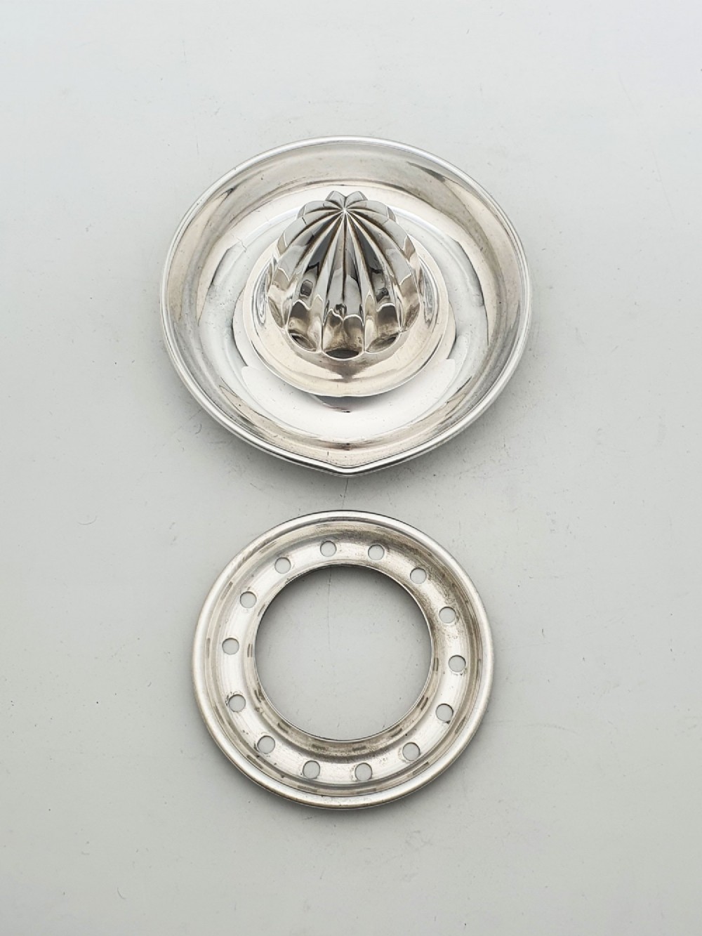 A silver-plated manual citrus squeezer featuring a central fluted reamer cone, a circular perforated strainer ring, and a shallow catch tray. View III