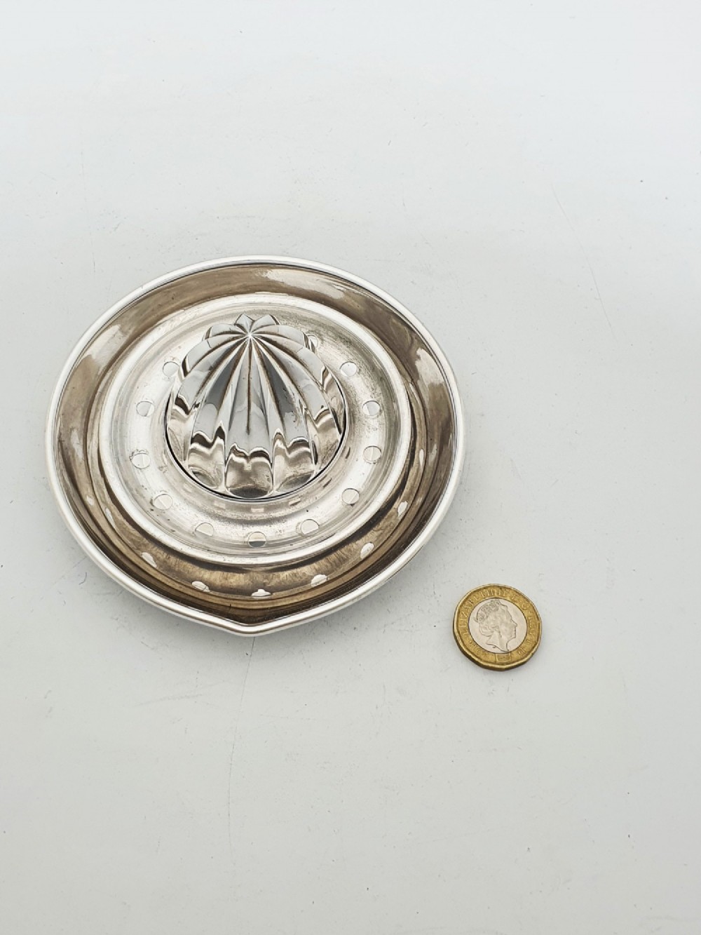 A silver-plated manual citrus squeezer featuring a central fluted reamer cone, a circular perforated strainer ring, and a shallow catch tray. View V
