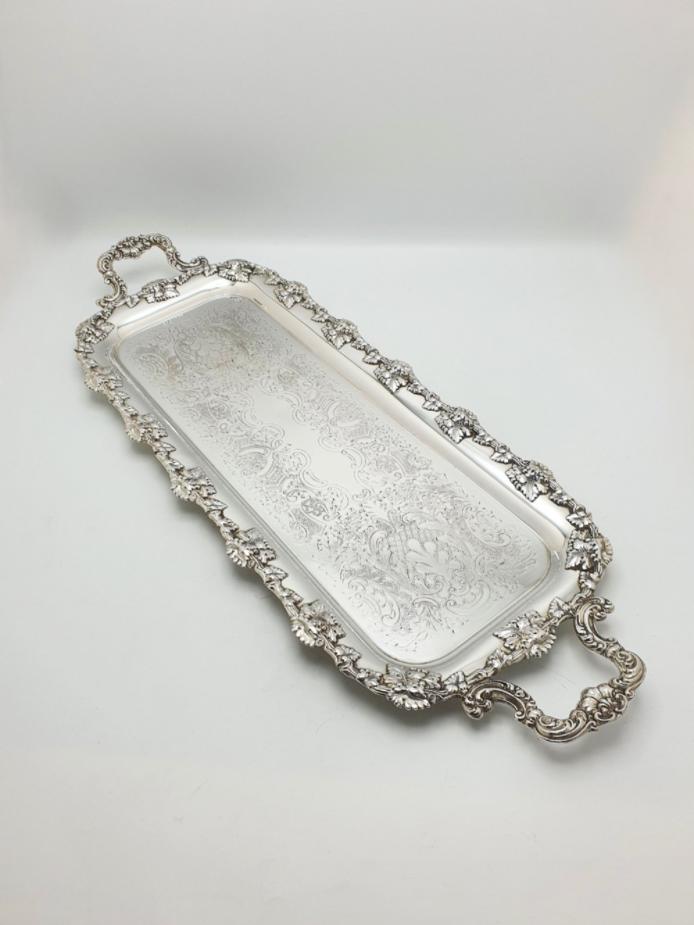 A long rectangular silver plated serving tray featuring an ornate grapevine motif rim, twin cast scroll handles, and an intricately chased floral central surface pattern.