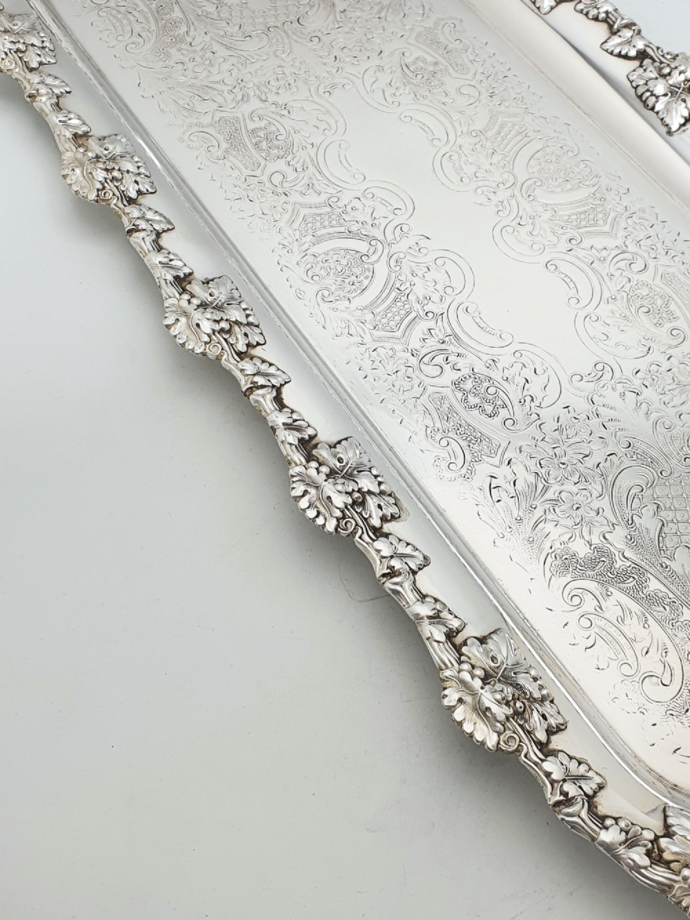 A long rectangular silver plated serving tray featuring an ornate grapevine motif rim, twin cast scroll handles, and an intricately chased floral central surface pattern. View V