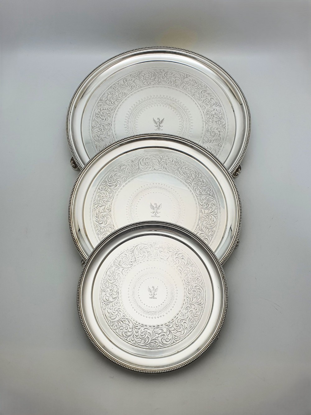 A set of three circular Victorian silver plated salvers arranged in graduated sizes, featuring beaded borders, detailed scroll engraving, and central crests.