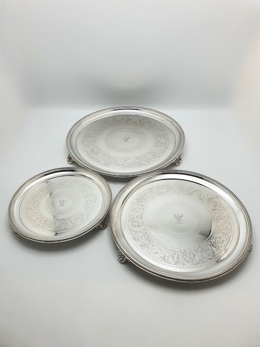 A set of three circular Victorian silver plated salvers arranged in graduated sizes, featuring beaded borders, detailed scroll engraving, and central crests. View II