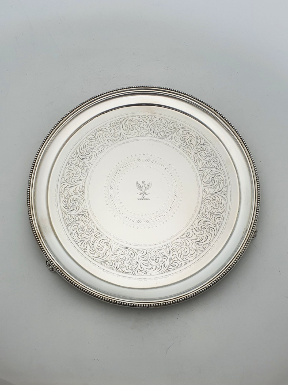 A set of three circular Victorian silver plated salvers arranged in graduated sizes, featuring beaded borders, detailed scroll engraving, and central crests. View V